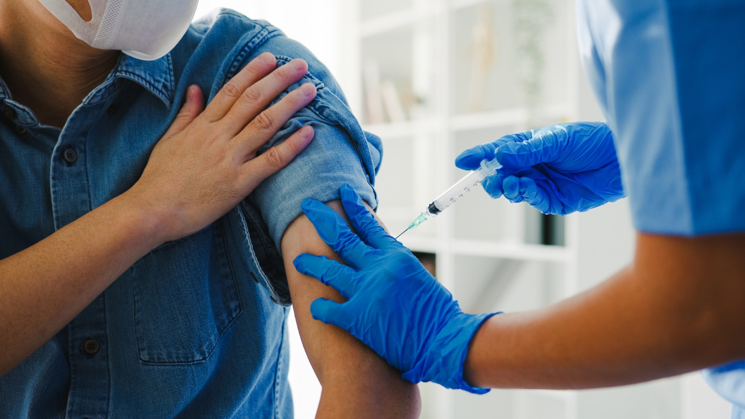 A person receives a vaccine injection in the upper arm from a healthcare worker wearing blue gloves.