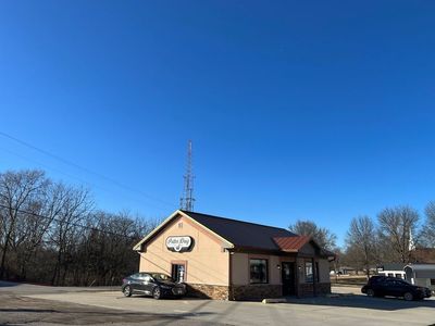 d29c4d74-d835-4c25-b077-c406e099998e_medium Small restaurant with a red roof and two parked cars under a clear blue sky, trees in the background.